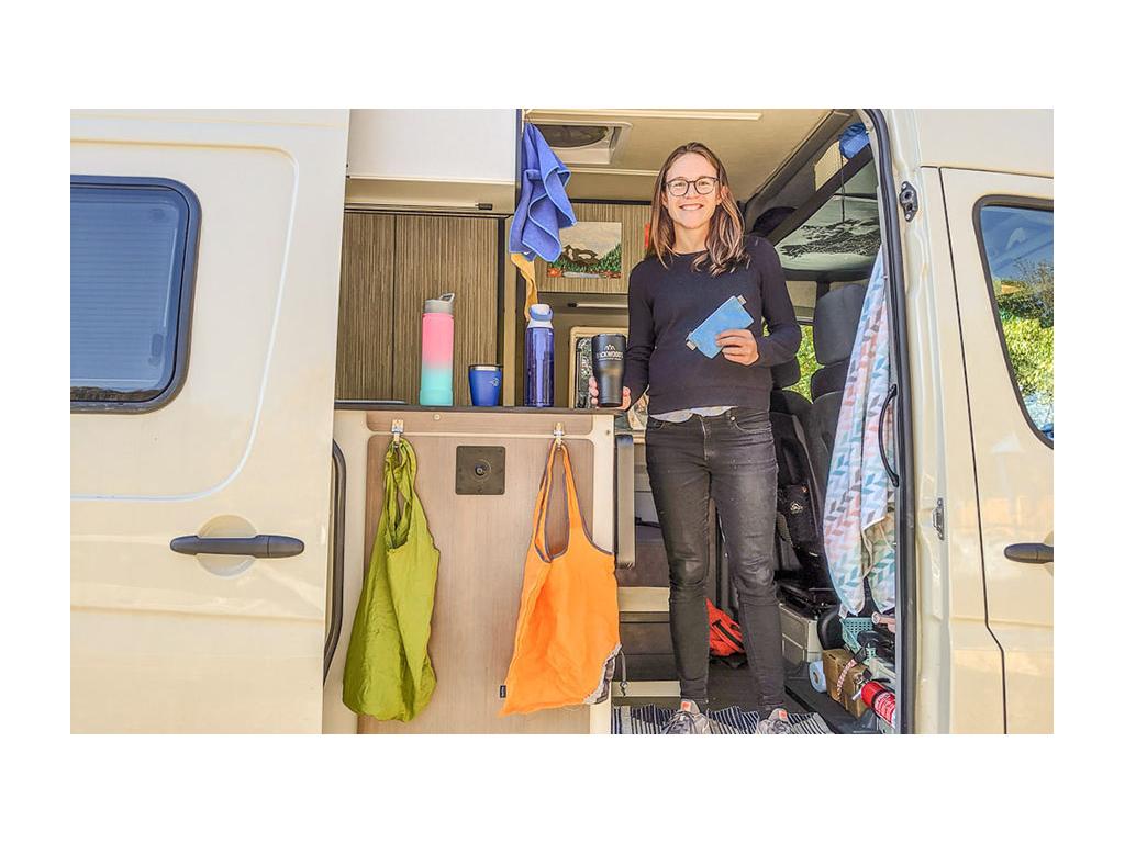 Kelly standing in Revel doorway with reusable water bottles, coffee mugs, and bags
