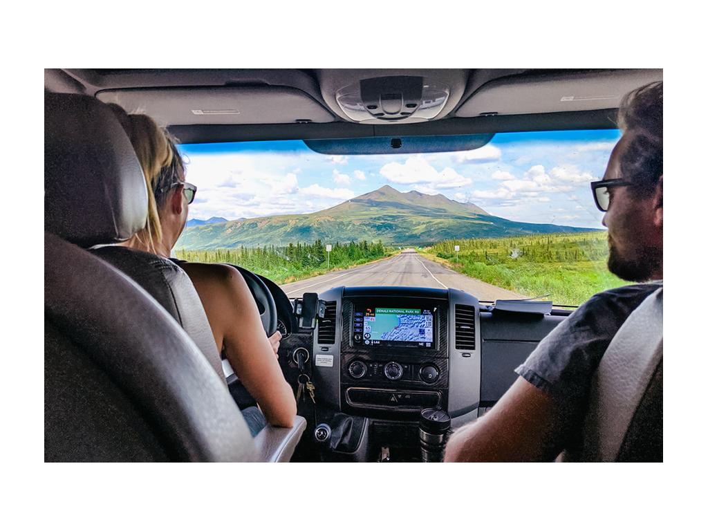 Photo from behind Katelyn driving with Howard in passenger seat .Looking out windshield to open road and mountains