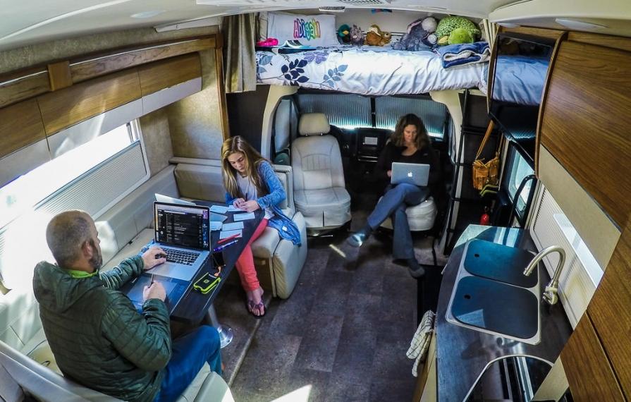 Peter and Abby sitting at dinette and Kathy sitting in passenger seat turned towards them. Peter and Kathy are on laptops and Abby is writing on note cards.