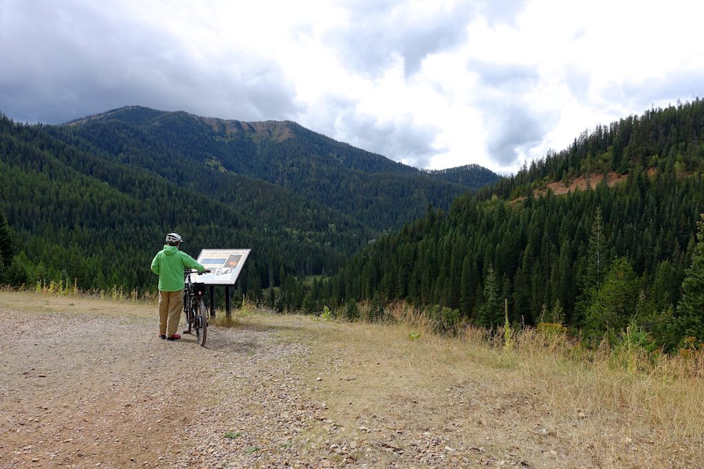Biker paused at a sign overlooking tree filled valley beneath the mountains.
