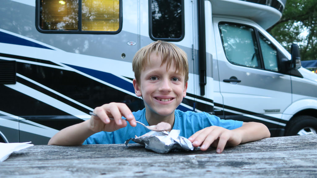 Child sitting at picnic table eating a snack with Winnebago View parked behind.