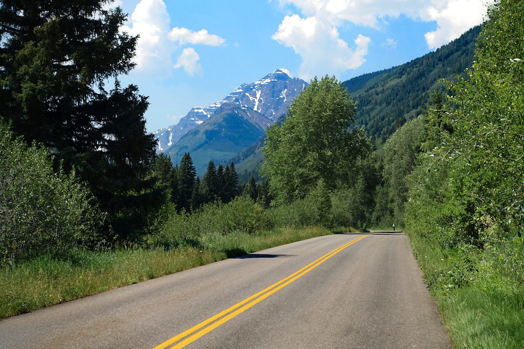 Paved road leading through the trees with mountains ahead.