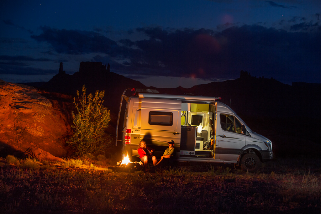 Peter and Kathy sitting by a campfire just outside their Winnebago Revel. 