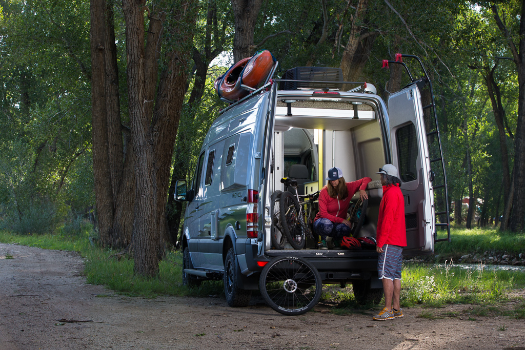 Man and woman at the back of the Winnebago Revel looking at their bikes that are stored.