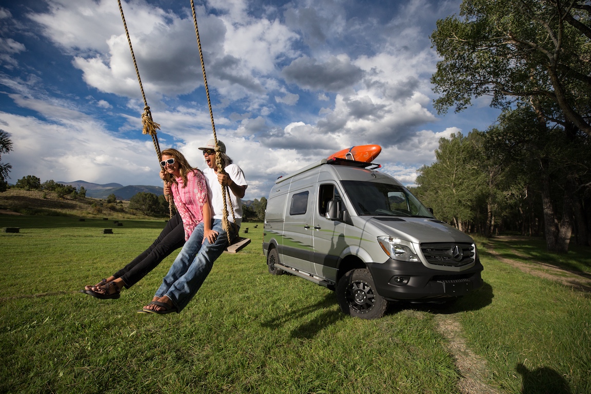 Winnebago Revel parked on the grass next to trees with a couple on a tree swing next to it.