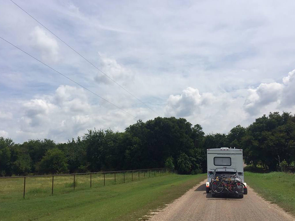 Motorhome with tow car attached driving down gravel road with field to the left and trees ahead.