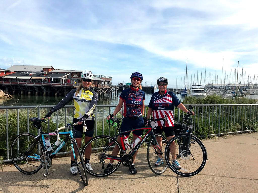 Three individuals standing next to their bikes with boats docked in the background