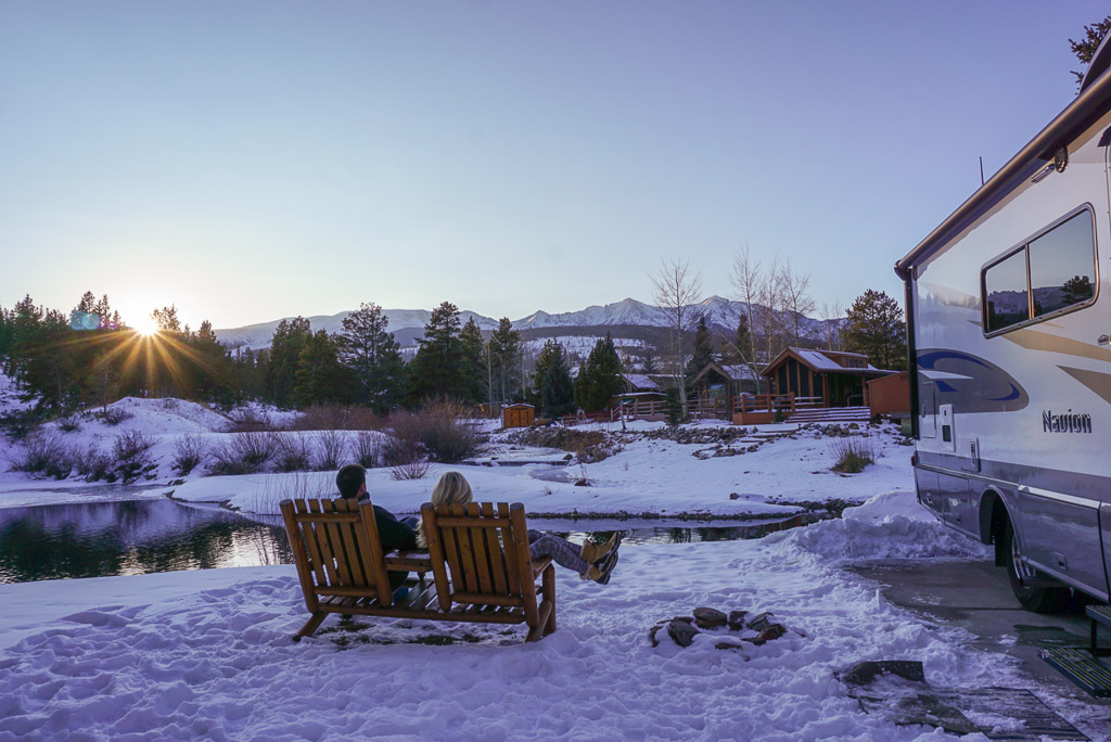 Couple sitting in chairs outside their Winnebago Navion looking out over a pond. Ground is snow covered.