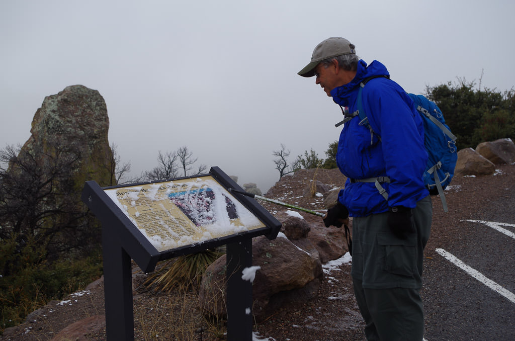 Man looking at snow covered sign with fog completely hiding scenery in front of him.