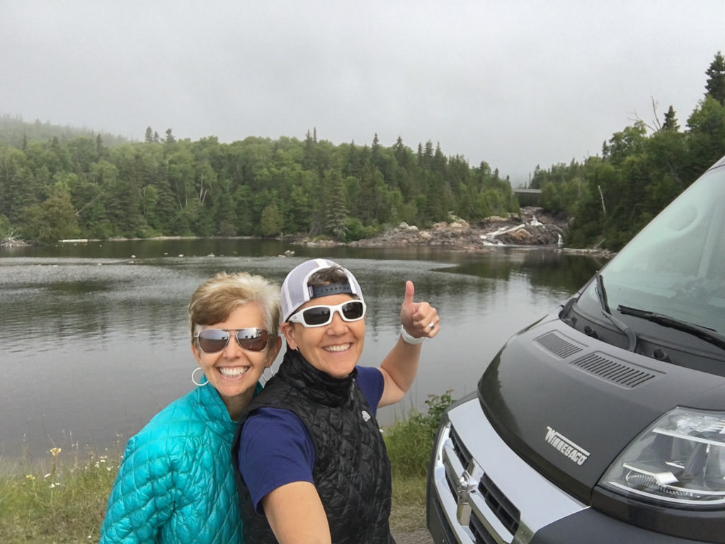 Noel and Chris standing next to their Winnebago Travato parked right next to the water.