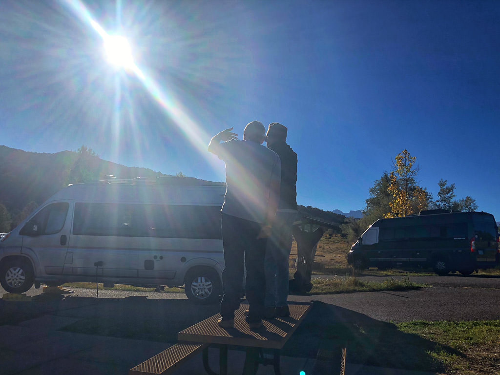 Two men standing on top of a picnic table looking across the way at something.
