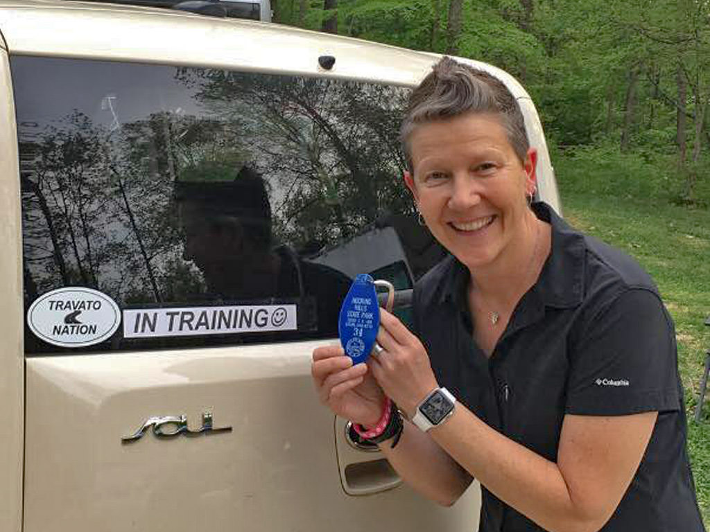 Woman standing behind vehicle with "Travato Nation" and "In Training" stickers on back.