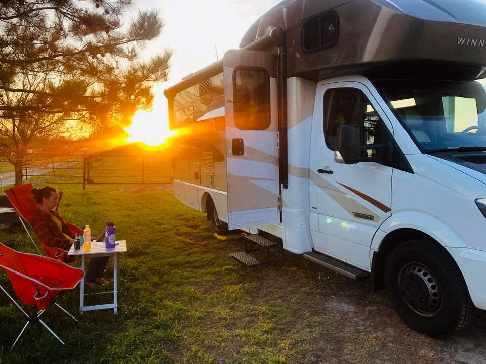 Woman sitting in camping chair outside Winnebago View with sun setting in the background.