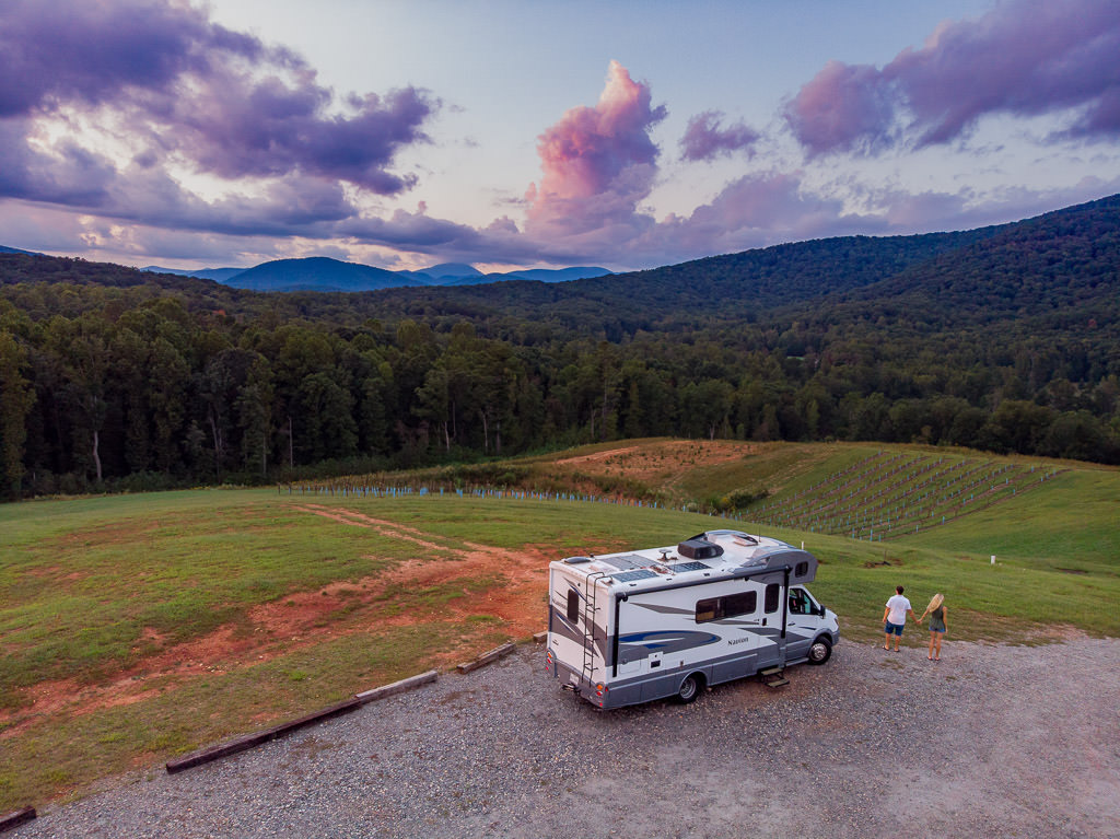 Couple holding hands standing outside their Winnebago Navion that is parked next to vines at Cottage Vineyard